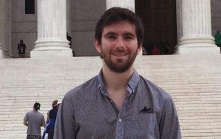 A young man in his 20s stands in front of a large building made of white marble with columns and stairs. The young man, whose name is David Whipple, is wearing a gray shirt and smiling warmly.