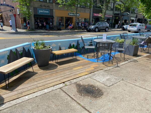 A parklet in Coolidge Corner, Brookline MA. It is outdoors and has several benches, tables, chairs, and green plants decorating it.