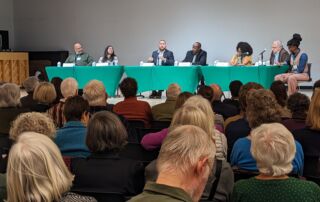 A crowd of people watches a panel discussion in an auditorium. The panelists are sitting side-by-side at a long table with a bright green tablecloth. They are engaged in conversation and eagerly sharing ideas.