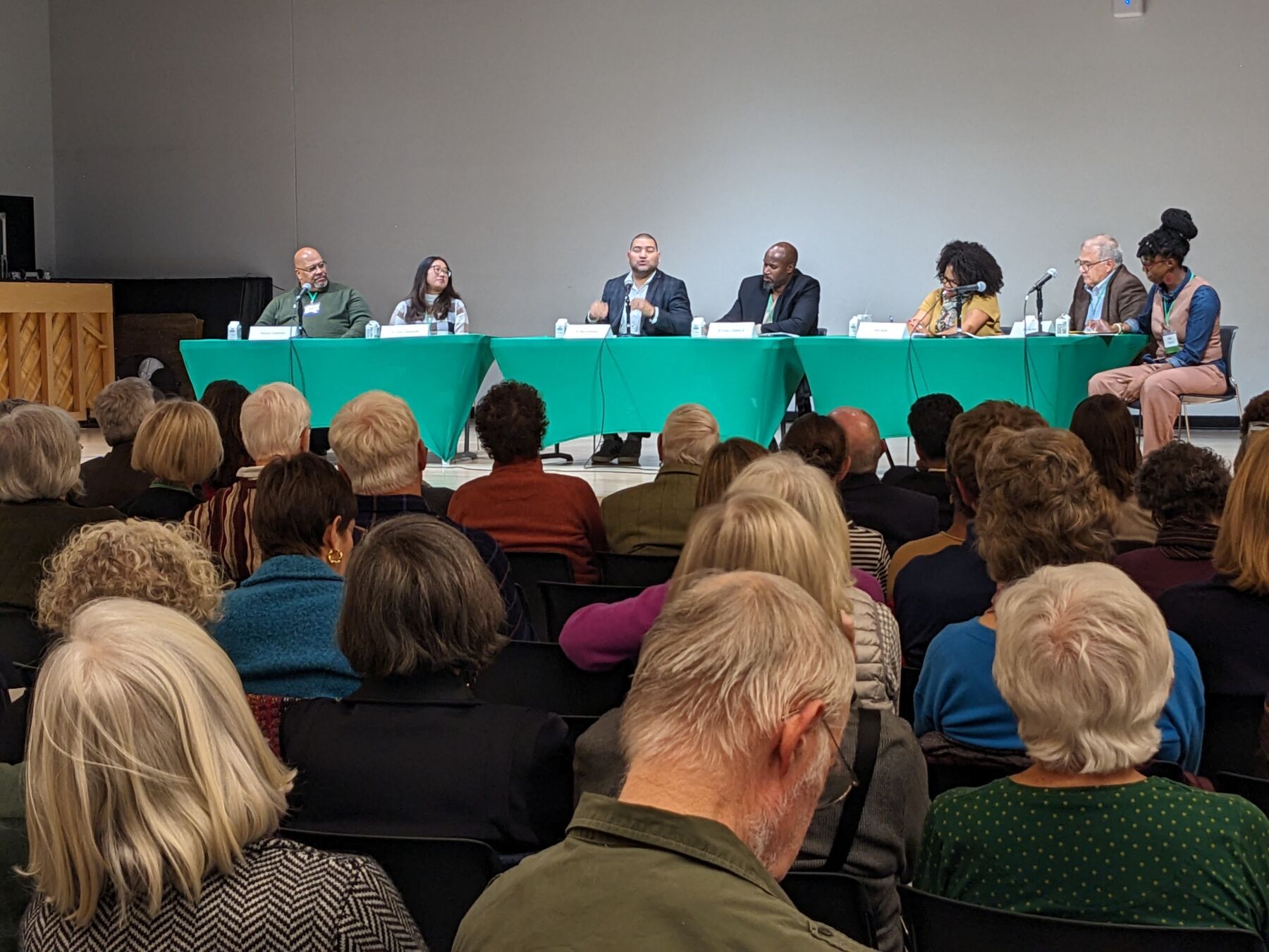 A crowd of people watches a panel discussion in an auditorium. The panelists are sitting side-by-side at a long table with a bright green tablecloth. They are engaged in conversation and eagerly sharing ideas.